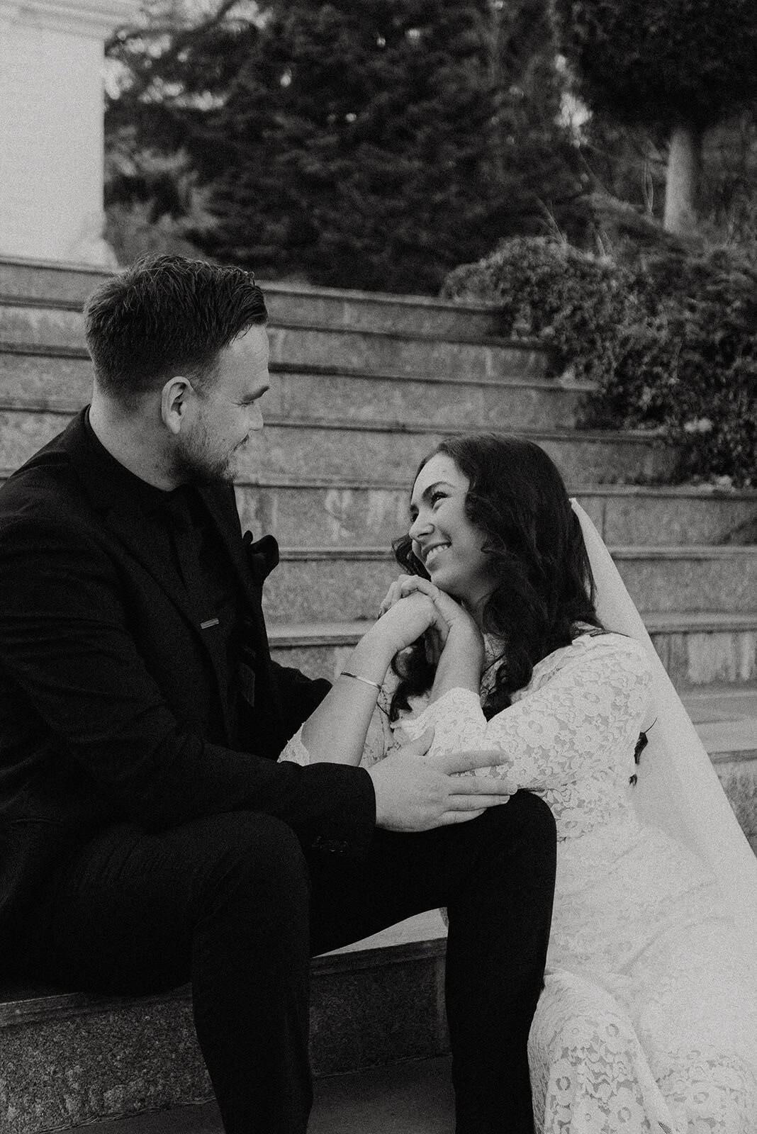 A woman in a wedding dress and a man in a suit sitting on outdoor stairs, smiling and sharing a mome.