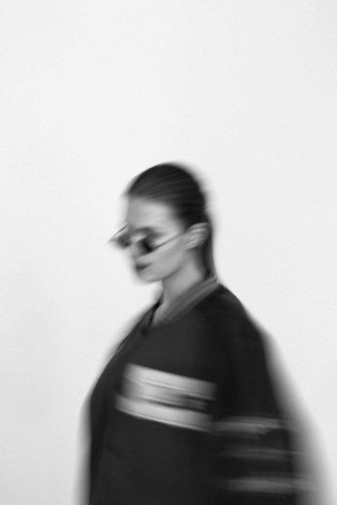 A woman wearing sunglasses and a black jacket, standing against a plain background.