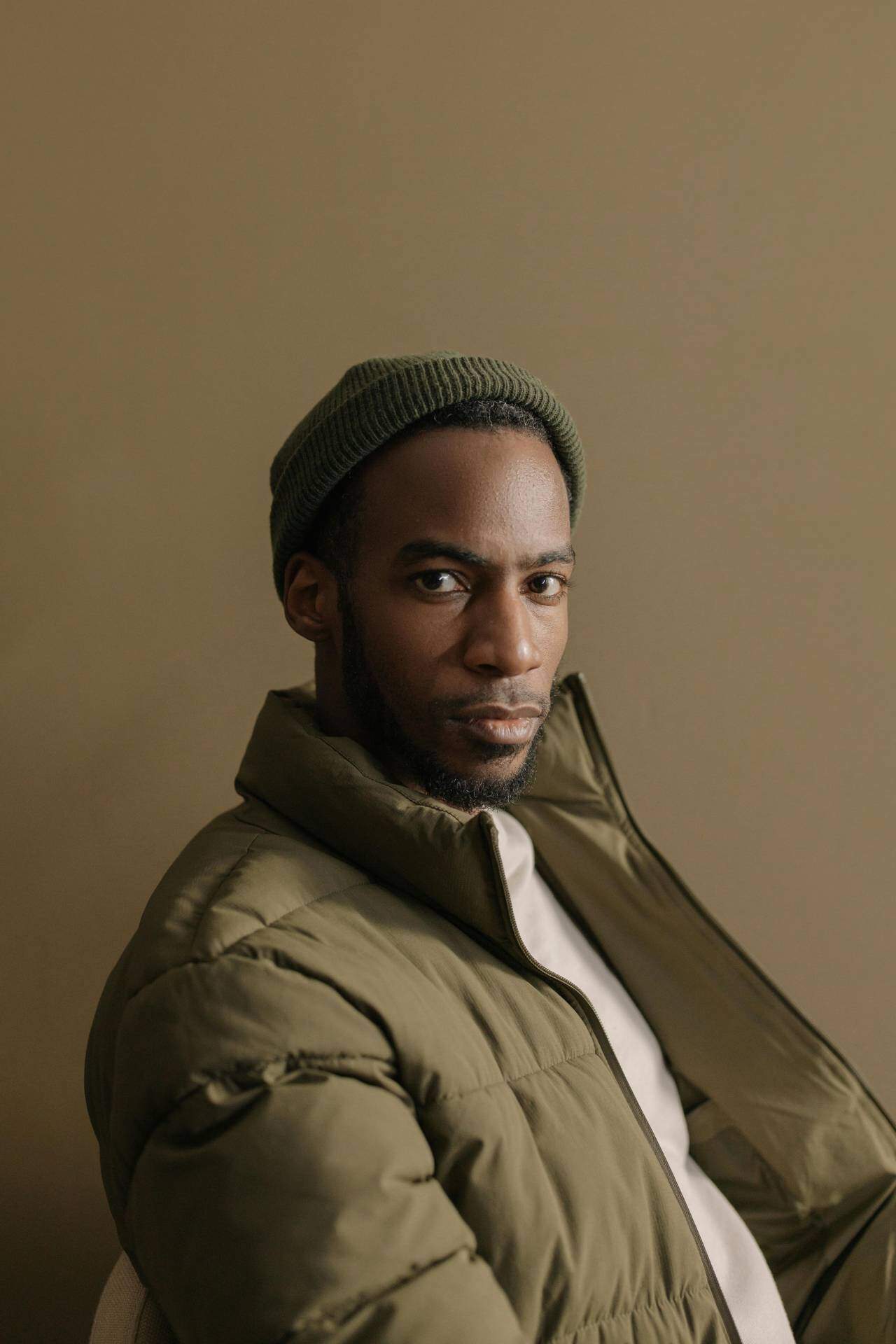 A woman with a serious expression wearing a green beanie and olive green jacket against a neutral ba.