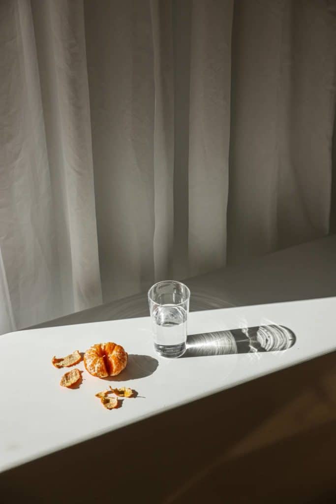A glass of water with orange peels on a white surface near a window with curtains.
